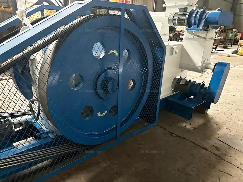 Large blue flywheel and drive belt system with a protective safety mesh on an industrial biomass briquette machine.