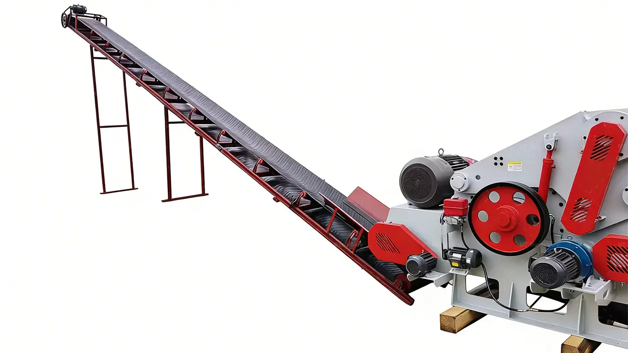 A detailed side view of a large industrial wood chipping system, focusing on the interface between the extensive, inclined feed conveyor and the grey-bodied drum chipper, which features prominent red pulleys and drive belts.