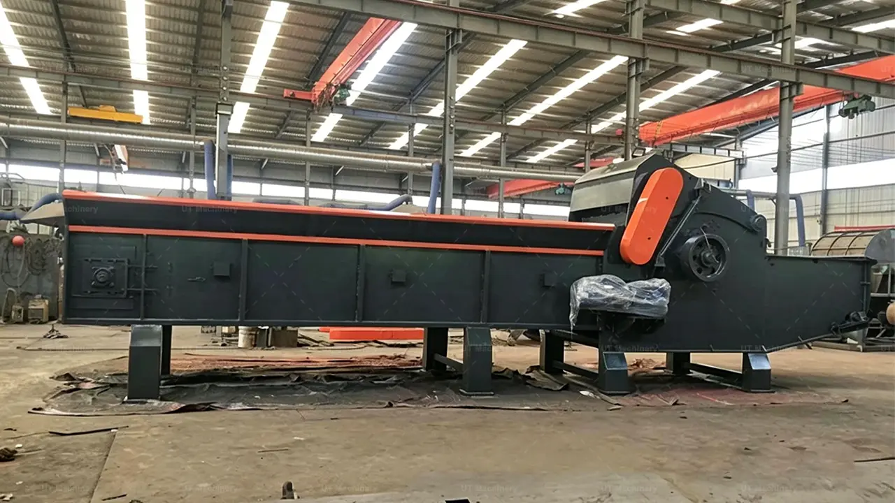 A long-distance, eye-level side profile shot of a massive, dark grey industrial wood chipper inside a well-lit workshop. The machine features a very long, low feeding trough on the left with an integrated chain-plate feeding system, leading to the large processing unit on the right.